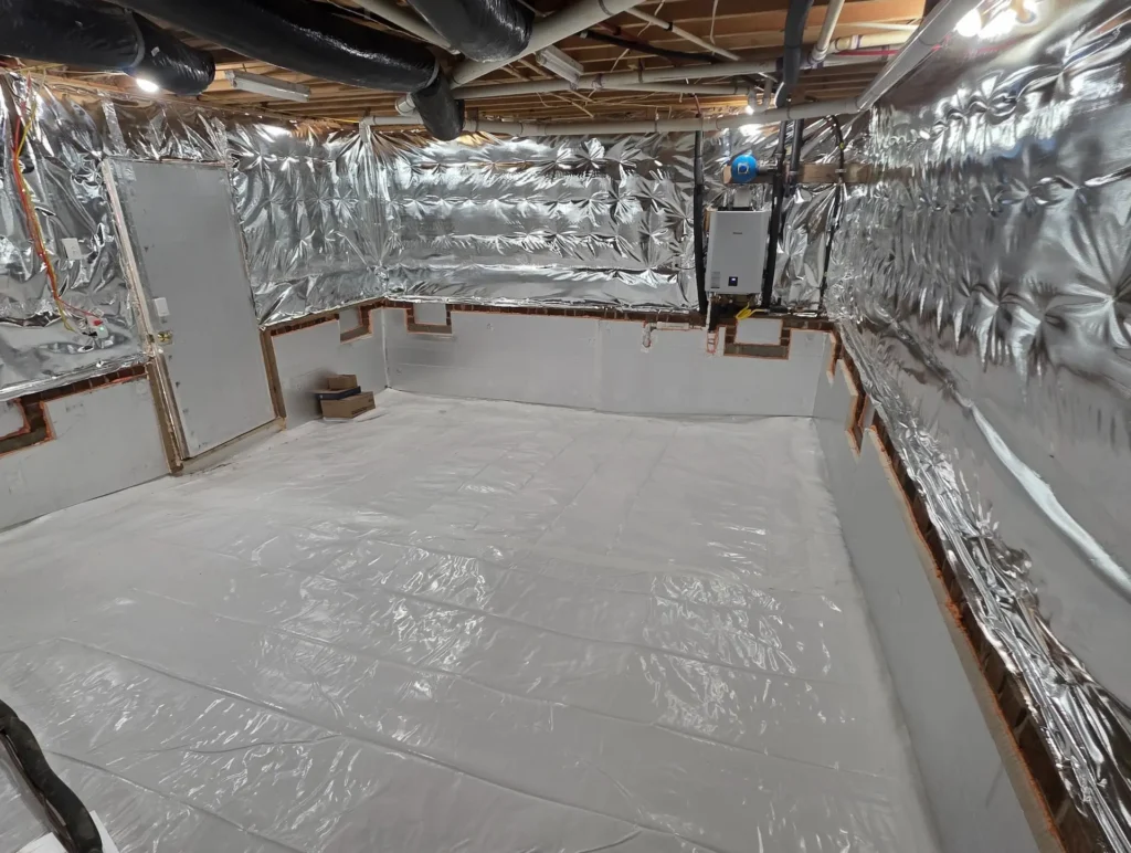 Picture of a brighly lit friendly healthy sealed crawlspace with bright white plastic on ground and hardboard insulation on lower wall and in and on pony wall is R19 spun insulation with a radiant barrier covering artistically fastened for appeal.